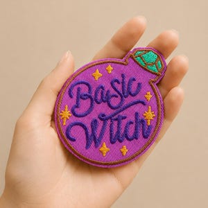 Può includere: Toppa ricamata a forma di pozione viola con la scritta "Basic Witch" in una sceneggiatura stilizzata. La toppa presenta accenti di stelle dorate e un dettaglio di gemma verde nella parte superiore. La toppa è tenuta in una mano.