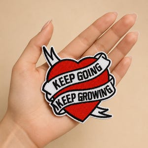 Könnte beinhalten: Ein roter herzförmiger Aufnäher mit einem weißen Banner mit der Aufschrift "KEEP GOING" und "KEEP GROWING" in schwarzem Text. Der Aufnäher hat eine schwarze Umrandung und wird in einer Hand gehalten.