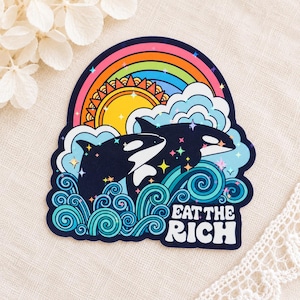 Op de afbeelding: Een gestanste sticker met twee zwart-witte orka's die door blauwe, wervelende oceaangolven springen. Boven de walvissen bevinden zich een heldergele zon, lichtblauwe wolken en een regenboog. De tekst EAT THE RICH is in het wit gedrukt.