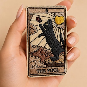 Può includere: Toppa ricamata raffigurante un gatto nero che afferra una farfalla, su uno sfondo di sole e montagne. Il disegno è incorniciato da un bordo marrone e include il testo "THE FOOL".