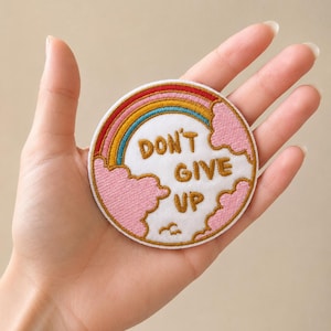 Könnte beinhalten: Runder, gestickter Aufnäher mit Regenbogen und dem Schriftzug "DON'T GIVE UP" in Weiß. Der Aufnäher hat einen goldenen Rand und rosa Wolkenakzente. Ein motivierendes Accessoire.