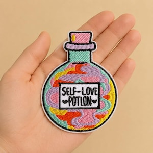 Könnte beinhalten: Gestickter Aufnäher in Form einer Zaubertrankflasche mit den Worten "SELF-LOVE POTION" in einem weißen Rechteck. Die Flasche hat einen rosa Verschluss, einen türkisfarbenen Hals und ein farbenfrohes Inneres mit Wirbeln aus Gelb, Orange, Rosa und Lila.