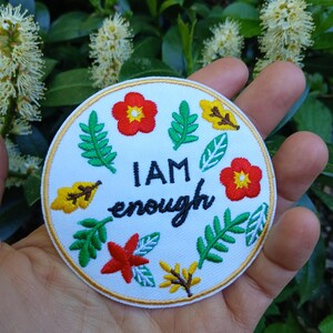 Op de afbeelding: Ronde, witte, geborduurde patch met de zwarte tekst "I AM enough". De patch is versierd met rode en gele bloemen, groene bladeren en een gouden rand. De patch wordt in een hand gehouden.