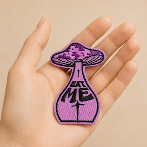 Op de afbeelding: Een paarse paddenstoelvormige patch met zwarte geborduurde details. De patch heeft de tekst "EAT ME" en een gestileerd ontwerp. De paddenstoelhoed is een donkerdere tint paars met een textuurpatroon.