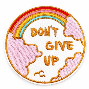 Può includere: Una toppa ricamata circolare con sfondo bianco e il testo arancione "DON'T GIVE UP". Il design include un arcobaleno colorato sopra il testo, circondato da nuvole rosa e dalla sagoma di un piccolo uccello arancione in basso.