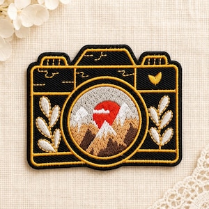 Op de afbeelding: Een zwarte geborduurde patch in de vorm van een camera met gouden omlijningen. De lens toont een landschap met bruine bergen, witte besneeuwde toppen en een grote rode zon. Witte bladachtige takjes versieren de zijkanten van de camerabody.