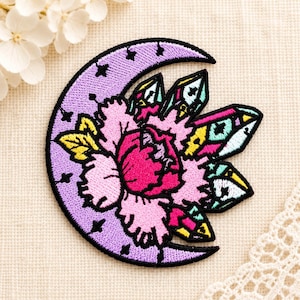 Könnte beinhalten: Ein bestickter Aufnäher mit einer lavendelfarbenen Mondsichel, die mit kleinen schwarzen Sternen verziert ist, einer großen rosa Blume in der Mitte und mehreren bunten Kristallen in Gelb, Türkis und Magenta mit schwarzen Umrandungen.