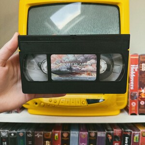 May include: A black VHS tape of the movie "Spirited Away" is held in front of a yellow television. The tape has a colorful label. Other VHS tapes are visible on a shelf below, including "The Holdovers" and "The Conjuring".