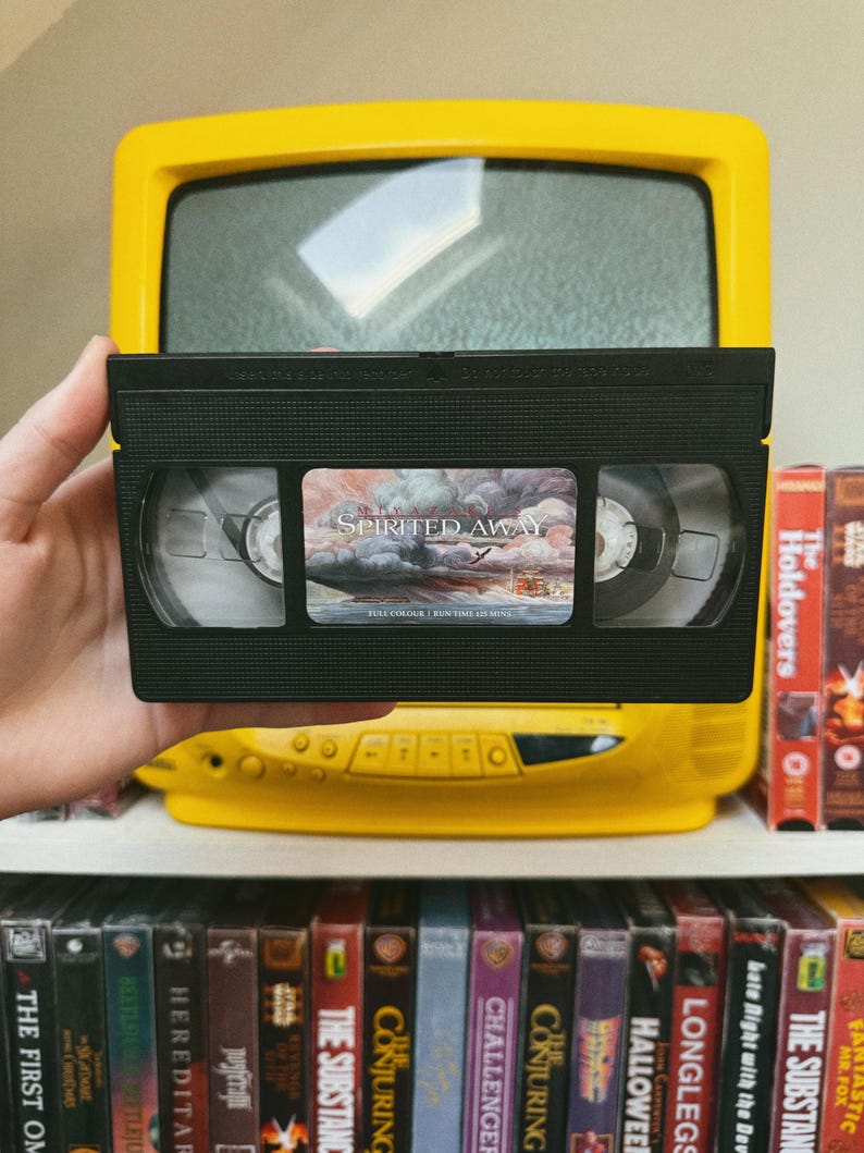 May include: A black VHS tape of the movie "Spirited Away" is held in front of a yellow television. The tape has a colorful label. Other VHS tapes are visible on a shelf below, including "The Holdovers" and "The Conjuring".