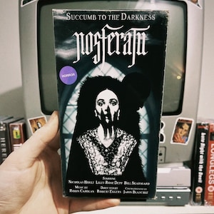 May include: A VHS tape for the horror movie "Nosferatu" starring Nicholas Hoult, Lilly-Rose Depp, and Bill Skarsgård. The movie is directed by Robert Eggers and the music is by Robin Carolan. The VHS tape has a black and white image of a woman with a shocked expression on her face.