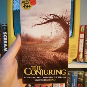 May include: A VHS tape of the horror film "The Conjuring" is held up in front of a vintage yellow television. The cover art depicts a haunting scene with a house, tree, and noose. The tape is marked "ADULTS ONLY 18+".