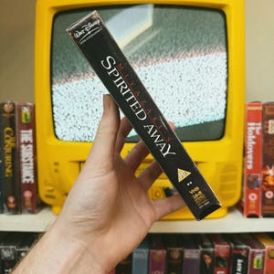 May include: A black VHS tape of the animated film "Spirited Away" is held in front of a yellow television. The tape's spine displays the film's title and the Walt Disney Home Entertainment logo. Other VHS tapes are visible on a shelf.