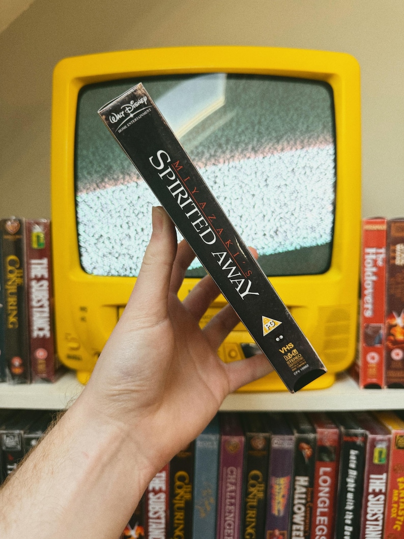 May include: A black VHS tape of the animated film "Spirited Away" is held in front of a yellow television. The tape's spine displays the film's title and the Walt Disney Home Entertainment logo. Other VHS tapes are visible on a shelf.