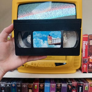 May include: A black VHS tape of "Howl's Moving Castle" held in front of a yellow television. The tape's label features an illustration of characters from the film. A shelf below displays various VHS tapes, including "The Holdovers" and "Fantastic Mr. Fox."