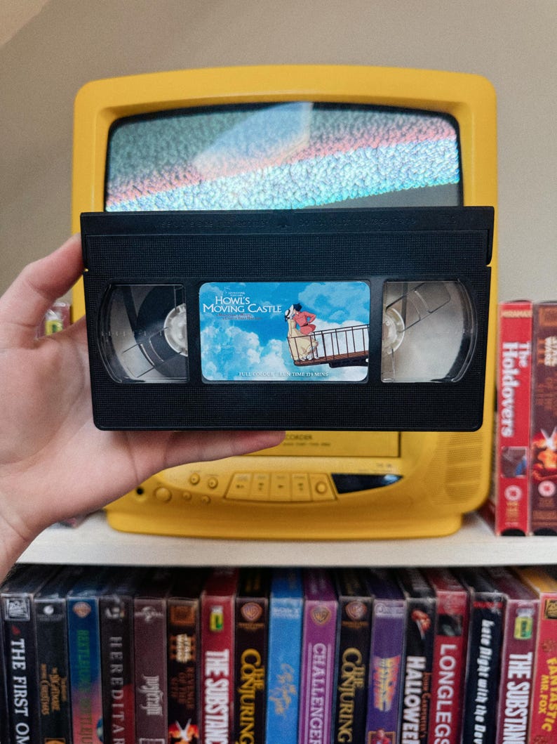 May include: A black VHS tape of "Howl's Moving Castle" held in front of a yellow television. The tape's label features an illustration of characters from the film. A shelf below displays various VHS tapes, including "The Holdovers" and "Fantastic Mr. Fox."