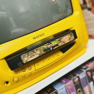 May include: A bright yellow Matsui television with a built-in VCR. The VCR has a tape inserted with a colorful image. The TV is on a white shelf with a collection of VHS tapes below.