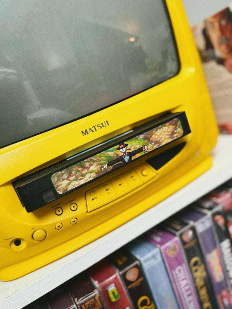 May include: A bright yellow Matsui television with a built-in VCR. The VCR has a tape inserted with a colorful image. The TV is on a white shelf with a collection of VHS tapes below.