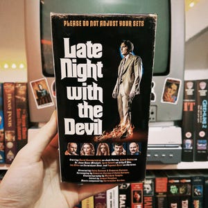 May include: A VHS tape case with a black background and white text that reads "Late Night with the Devil". The case features a man in a suit standing in front of a fiery background. The text "Please do not adjust your sets" is at the top of the case.