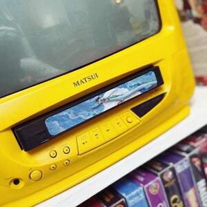 May include: A bright yellow Matsui TV with a built-in VCR. The VCR is playing a video. Below the TV are several VHS tapes on a shelf. The TV has buttons for volume, channel, and playback.
