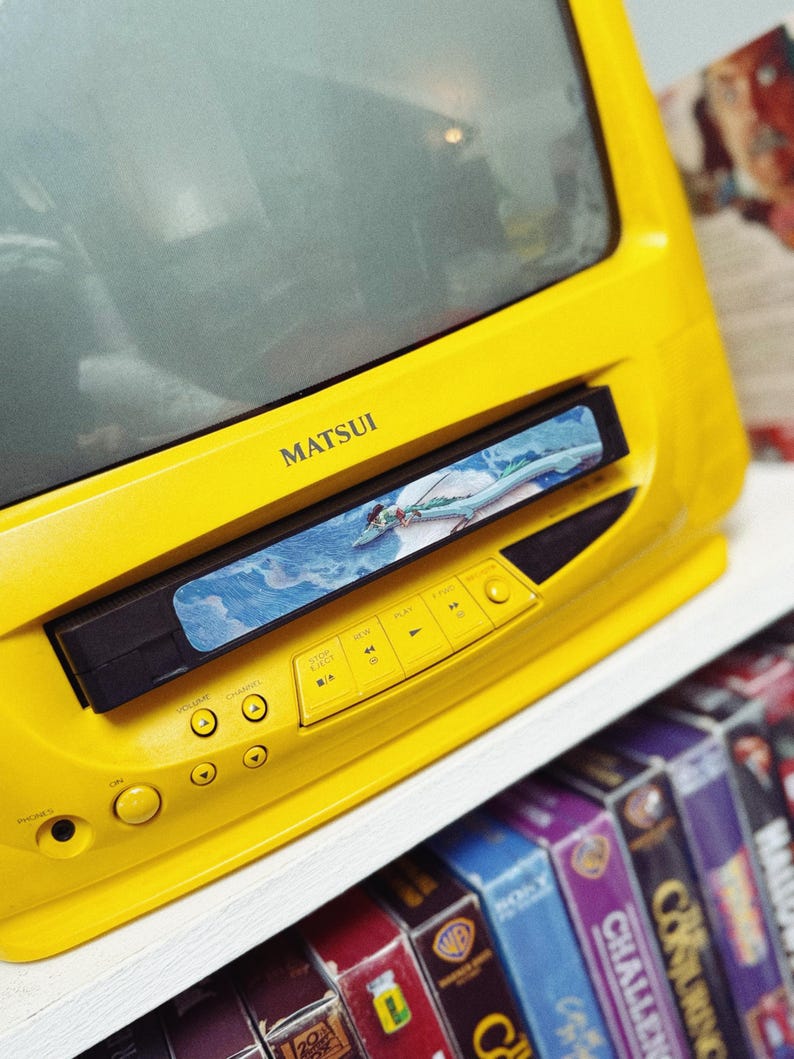 May include: A bright yellow Matsui TV with a built-in VCR. The VCR is playing a video. Below the TV are several VHS tapes on a shelf. The TV has buttons for volume, channel, and playback.