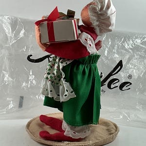 Pu&ograve; includere: Una statuina natalizia di una bambola che tiene una scatola regalo. La bambola indossa un abito rosso e verde con un cappello e un grembiule bianchi. La scatola regalo &egrave; bianca con un nastro rosso. La statuina &egrave; su una base rotonda e beige.