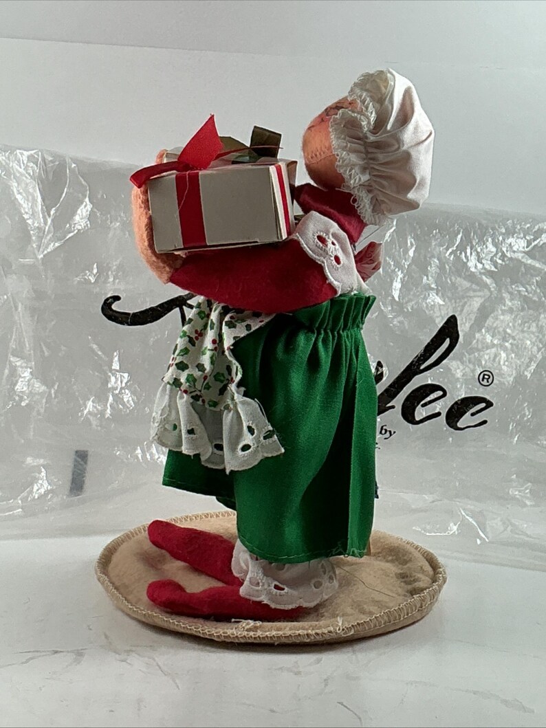 Pu&ograve; includere: Una statuina natalizia di una bambola che tiene una scatola regalo. La bambola indossa un abito rosso e verde con un cappello e un grembiule bianchi. La scatola regalo &egrave; bianca con un nastro rosso. La statuina &egrave; su una base rotonda e beige.