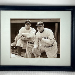 May include: Framed black and white photograph featuring two baseball players in vintage uniforms, leaning on bats. The photo is in a dark blue frame with a white matting, capturing a moment in baseball history.