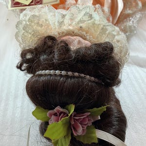 May include: A vintage doll from the Anna Collection, named "Cathryn." The doll has dark brown hair styled with a pearl headband and a floral accent. She wears a peach-colored dress with lace trim. A tag with the doll's name is visible.