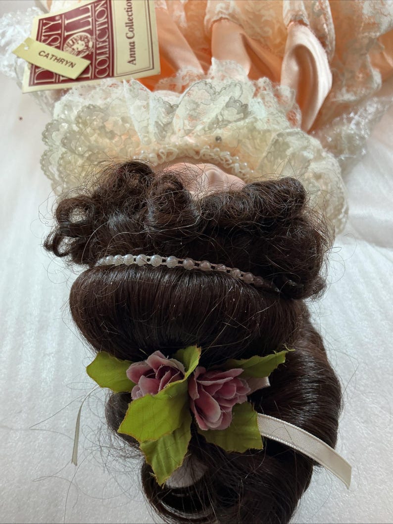 May include: A vintage doll from the Anna Collection, named "Cathryn." The doll has dark brown hair styled with a pearl headband and a floral accent. She wears a peach-colored dress with lace trim. A tag with the doll's name is visible.
