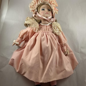 Könnte beinhalten: Eine Vintage-Porzellanpuppe in einem rosa Kleid und einer passenden Haube mit Spitzenbesatz. Die Puppe hat blondes Haar, blaue Augen und trägt braune Schuhe. Das Kleid hat lange Ärmel und ein gefaltetes Oberteil. Die Puppengröße beträgt ca. 30 cm.