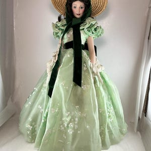 Puede incluir: Muñeca de porcelana con un vestido verde claro con detalles florales blancos, un sombrero de paja y una faja de terciopelo verde oscuro. La muñeca tiene la cara pintada y lleva una bufanda y guantes verdes a juego. El vestido de la muñeca tiene una falda amplia.