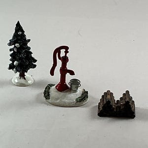 May include: Miniature Christmas village figurines. Includes a dark green Christmas tree with white accents, a red water pump with a bucket, and a stack of brown logs. The figurines are on a white base.