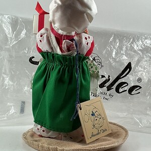 Pu&ograve; includere: Una bambola Annalee vintage, con un cappuccio bianco, un vestito rosso e bianco e una gonna verde. La bambola tiene in mano una piccola scatola regalo con un nastro rosso. La bambola &egrave; su una base rotonda e beige. Un'etichetta con il logo Annalee Dolls &egrave; attaccata.