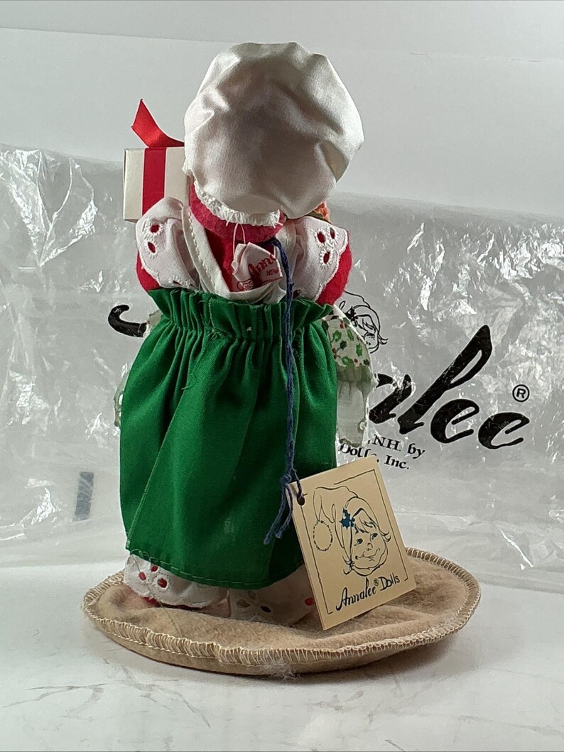 Pu&ograve; includere: Una bambola Annalee vintage, con un cappuccio bianco, un vestito rosso e bianco e una gonna verde. La bambola tiene in mano una piccola scatola regalo con un nastro rosso. La bambola &egrave; su una base rotonda e beige. Un'etichetta con il logo Annalee Dolls &egrave; attaccata.