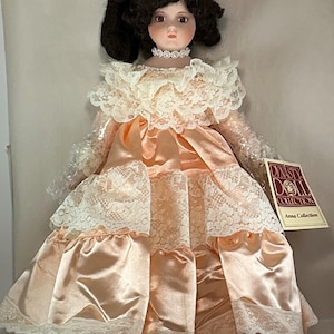 May include: A porcelain doll from the Dynasty Doll Collection, Anna Collection. The doll has dark hair styled in pigtails, a choker, and a peach-colored satin dress with white lace trim. The doll is wearing a peach-colored satin dress with white lace trim.