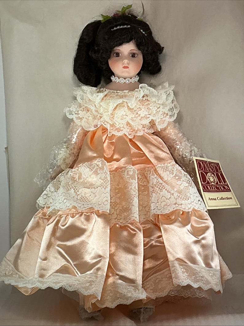May include: A porcelain doll from the Dynasty Doll Collection, Anna Collection. The doll has dark hair styled in pigtails, a choker, and a peach-colored satin dress with white lace trim. The doll is wearing a peach-colored satin dress with white lace trim.