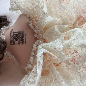 May include: A Dynasty Doll Collection doll with brown hair and a pearl necklace. The doll is wearing a peach-colored dress with a white lace collar. The doll's neck has the Dynasty Doll Collection logo.