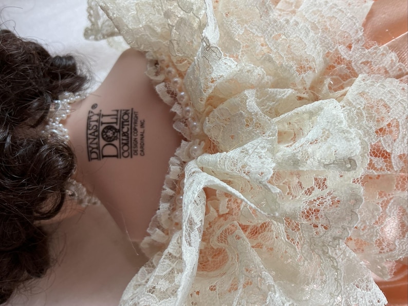 May include: A Dynasty Doll Collection doll with brown hair and a pearl necklace. The doll is wearing a peach-colored dress with a white lace collar. The doll's neck has the Dynasty Doll Collection logo.