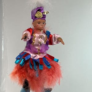May include: A hanging monkey doll ornament wearing a purple hat with a gold rose and white feathers. The doll features a pink satin jacket with gold trim, a skirt made of layered red and blue ribbons, and a fluffy coral feather skirt with blue shoes.