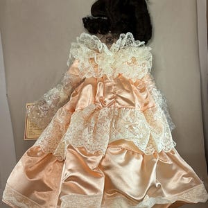 May include: A vintage doll with dark hair, wearing a peach-colored satin dress with white lace trim. The dress has a layered design and a ruffled collar. The doll is in its original packaging.