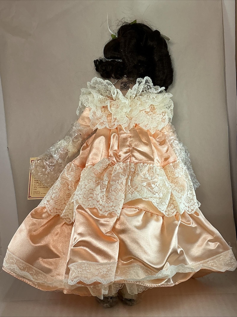 May include: A vintage doll with dark hair, wearing a peach-colored satin dress with white lace trim. The dress has a layered design and a ruffled collar. The doll is in its original packaging.