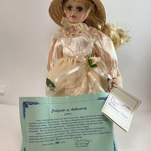 Può includere: Una bambola in porcellana con capelli biondi e occhi azzurri, che indossa un abito beige con pizzi, nastri e un cappello di paglia. Include un certificato di autenticità Duck House Heirloom Dolls, con il nome Patsy e il numero di serie 1084/5000.
