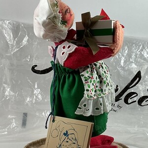 Pu&ograve; includere: Una bambola Annalee a tema natalizio, con un vestito verde, scarpe rosse e un cappello bianco. La bambola tiene in mano una scatola regalo con un nastro verde. &Egrave; presente un'etichetta Annalee Dolls.