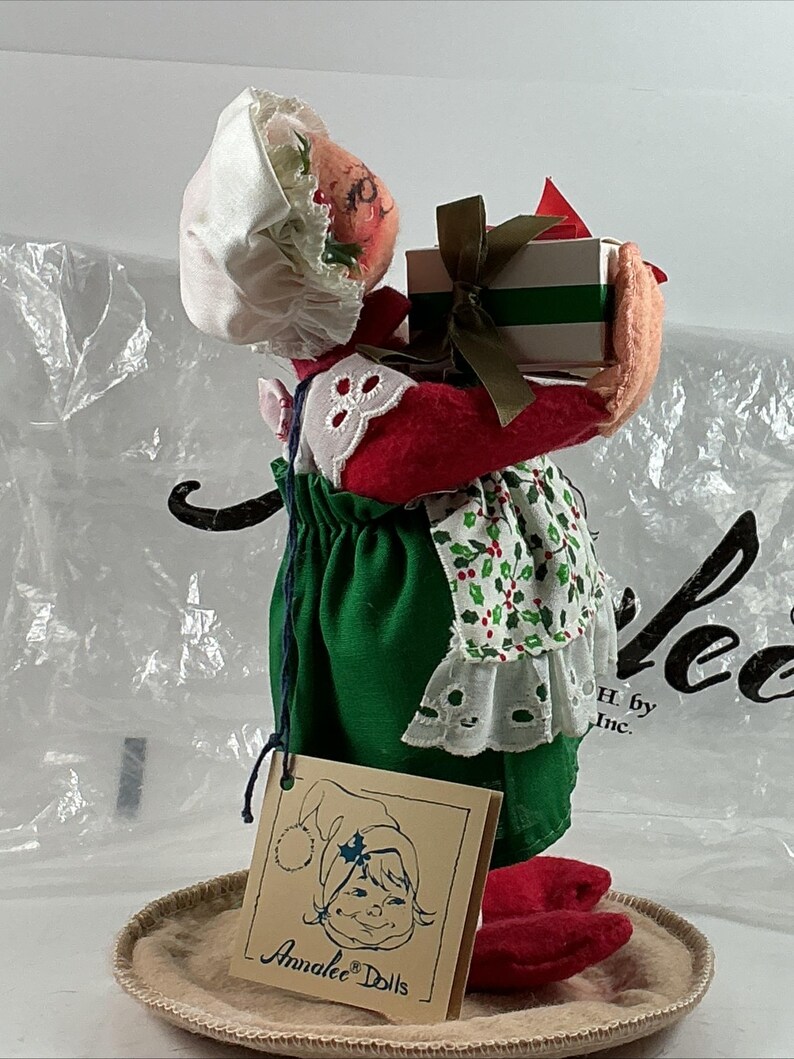 Pu&ograve; includere: Una bambola Annalee a tema natalizio, con un vestito verde, scarpe rosse e un cappello bianco. La bambola tiene in mano una scatola regalo con un nastro verde. &Egrave; presente un'etichetta Annalee Dolls.