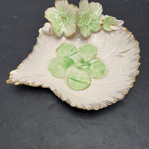 Op de afbeelding: Een wit porseleinen bladvormig schaaltje met een gouden rand en groene bloemmotieven. Het schaaltje heeft twee reliëfbloemen en een klavertje vier.