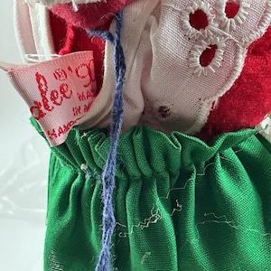 Pu&ograve; includere: Primo piano di abiti per bambole vintage. L'outfit della bambola include una gonna verde, un top rosso e bianco e un bordo in pizzo bianco. &Egrave; visibile un filo blu, insieme a un'etichetta rosa con "alee's".