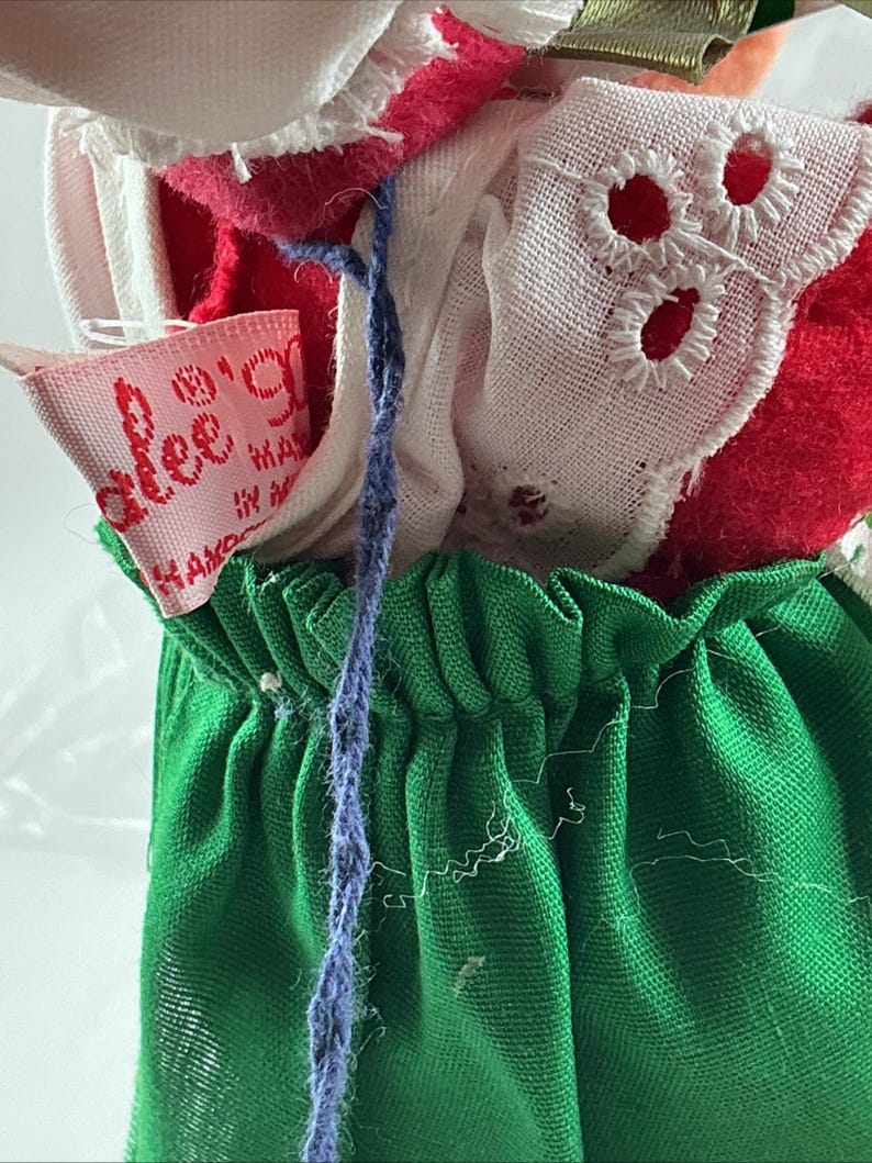 Pu&ograve; includere: Primo piano di abiti per bambole vintage. L'outfit della bambola include una gonna verde, un top rosso e bianco e un bordo in pizzo bianco. &Egrave; visibile un filo blu, insieme a un'etichetta rosa con "alee's".
