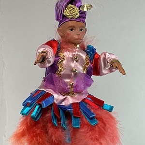 May include: A monkey doll wearing a purple turban with a gold rose, a pink and purple jacket with gold trim, and a skirt made of red and blue ribbons and orange feathers. The doll has light blue shoes and a white feather plume on the head.