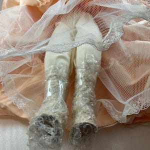 May include: A doll's legs and feet are visible, clad in off-white fabric and delicate lace-trimmed boots. The doll's legs are covered in bubble wrap. The doll is wearing a peach-colored dress with a white net overlay.