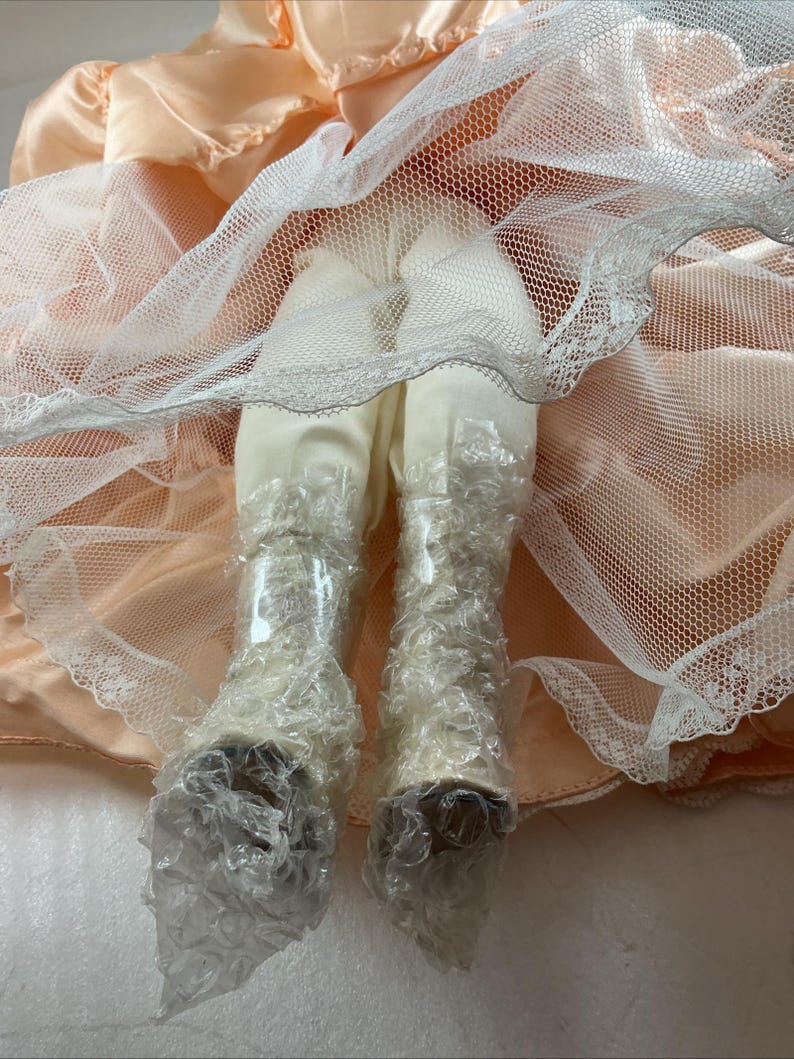 May include: A doll's legs and feet are visible, clad in off-white fabric and delicate lace-trimmed boots. The doll's legs are covered in bubble wrap. The doll is wearing a peach-colored dress with a white net overlay.